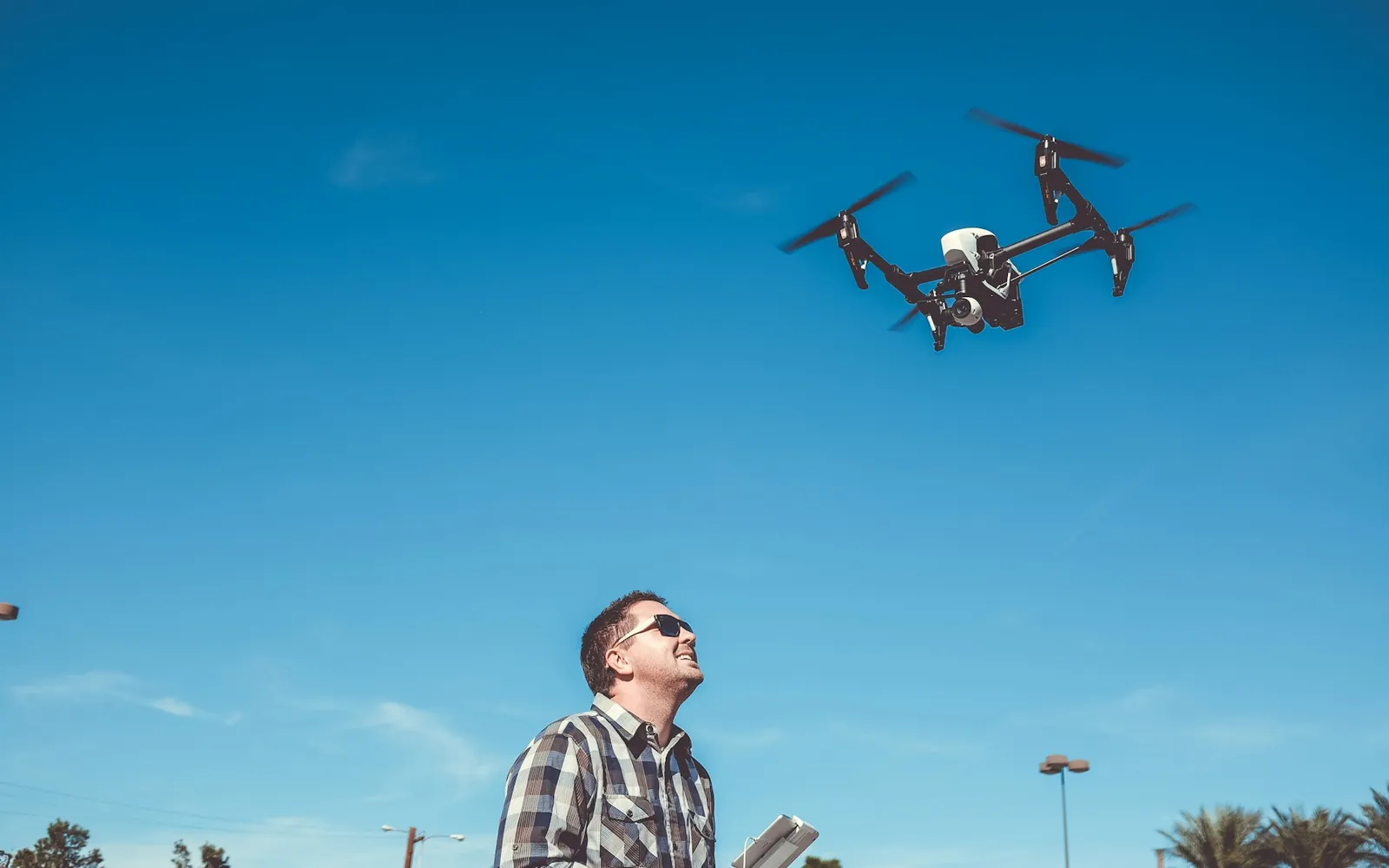 A remote pilot flying a quadcopter drone against a clear blue sky.